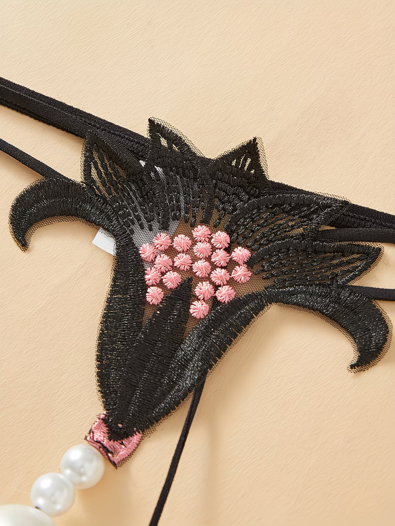 Bikinisexzy black lace embroidered pearl massage thong front view on beige background with pink roses